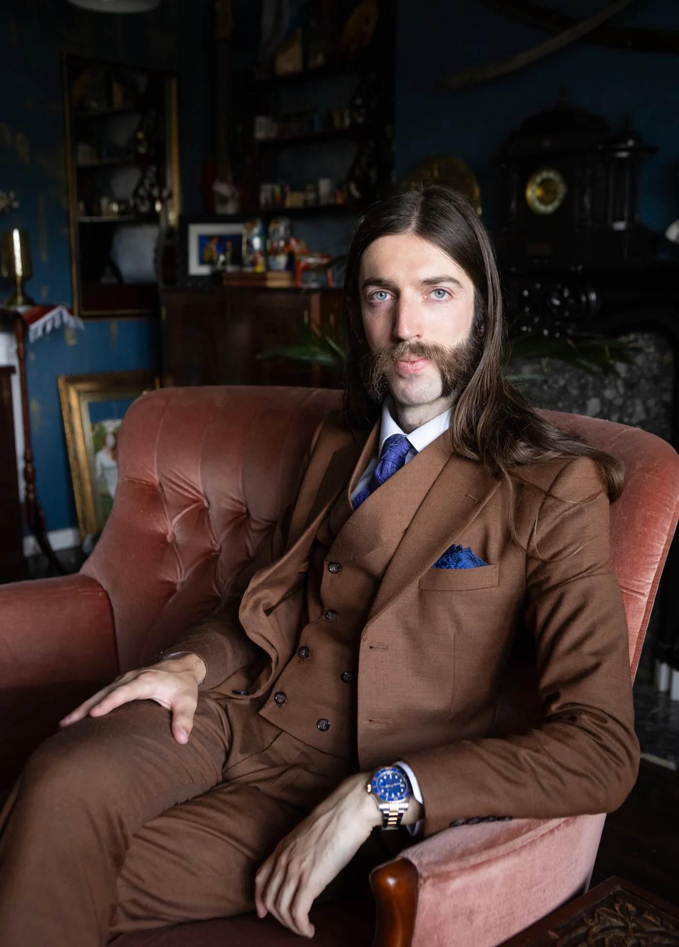 Stewart Vickers in a brown three-piece suit and blue paisley tie, seated in a pink velvet armchair in a blue room hung with pictures
