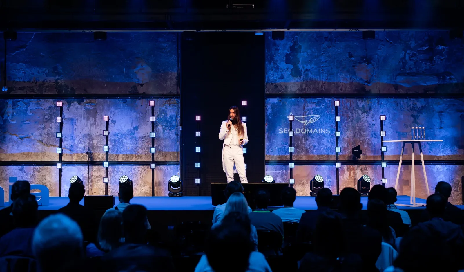 Stewart Vickers alone on a lit stage in a white suit at SEO Estonia in Tallinn, the audience silhouetted in the foreground