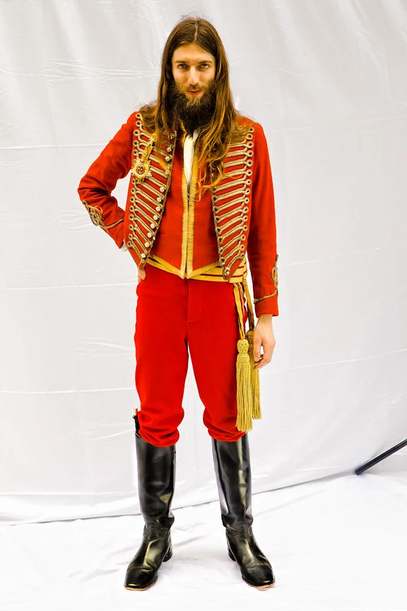 Studio portrait of Stewart Vickers in a red frogged hussar jacket, bullion sash and riding boots, against a plain backdrop